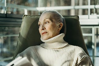 An older woman with grey hair pulled back in a ponytail wears a beige turtleneck sweater and glances sideways while sitting in an olive green chair.