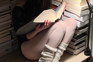 A ballerina, dressed in a dark leotard, with sheer white stockings. She wears off-white leg warmers, just over her ankle and the heel of her ballet shoes. She sits in a corner, with stacks of books surrounding her, filling the frame. We can’t see the spines of the books, just the pages. They’re all thick, holding, eluding to millions of words. Her head is buried deep in a huge tome as her hands cradle the book gently, dark brown hair obscuring her face. A white lamp illuminates the scene.