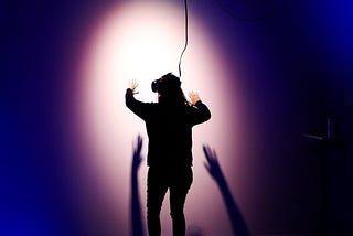 Person with a Virtual Reality headset gesturing in front of a wall, illuminated by a spotlight.