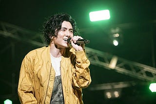 Fujii Kaze in a yellow jacket, singing into a mic, with a green light in the background.