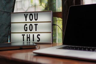 A laptop sitting on a desk with a sign beside it stating “You Got This”