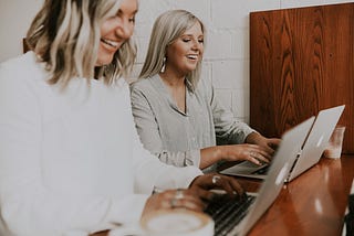 Two women blog at a desk with coffee.