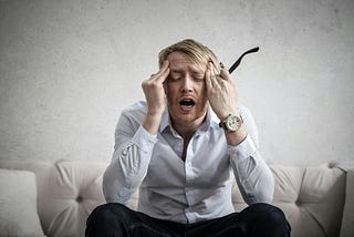 A young professional man sitting on a sofa, feeling pressure, taking his glasses in his left hand.