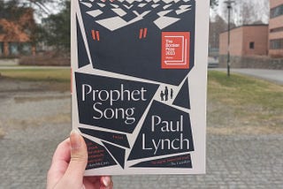 Fariza’s hand holding a copy of Prophet Song with trees in the background and red brick buildings to the left and right side of the picture. There is pavement and patch of dry grass in the lower half of the picture.