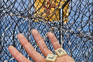 Prison, barbed wire, chain link fence, captive, yellow flag, in the foreground is an open hand holding Scrabble tiles which spell out the word Help