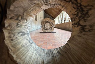 The image shows a view through a hollowed-out tree trunk, focusing on a cross-section of another tree trunk placed on a stand in the background. The scene is set in a corridor with stone walls and a tiled floor, featuring a row of columns and a slanted wooden roof. The perspective creates a tunnel-like effect, drawing attention to the intricate patterns and textures of the tree trunks.