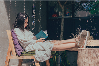 A woman reading a book sitting on a wooden chair with her feet up