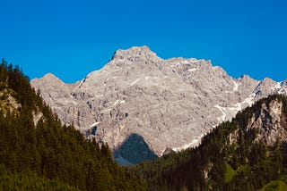 A beautiful bare-rock mountain with a few thin strips and pockets of remaining snow (the picture was taken in the height of summer) towering impressively into a deep blue sky. In the foreground the picture is framed by two steep slopes, one going left and the other right, covered in conifer forests.