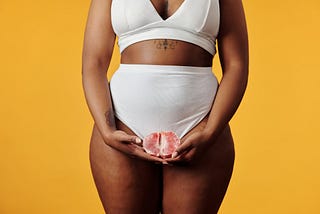 black woman in plain under garments holding a grapefruit in front of her privates