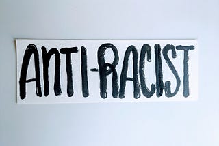 Hand lettering of “Anti-racist” in all capitalized letters painted in black ink on white strip of paper.