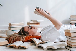 White, blonde woman lying on the floor, her head cushioned by a pillow, as she reads a book. She is surrounded by piles of books.