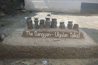 A traditional cooking setup from a tribal village with “The Sangyan: Ulgulan Talks!” written over it on the smaller platform and below the earthen chimney. The setup has eight small earthen chimneys over a small platform that settles over a larger platform all made up of soil and other natural ingredients and are brown in colour.