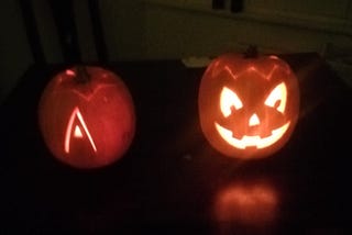 Two pumpkins, one a traditional jack-o-lantern, the other with the letter A carved into the front, both lit up from inside against a dark background