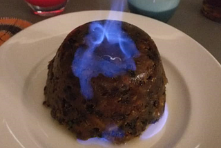 A Christmas pudding sits on a white plate with a blue flame rising out of the top.