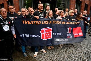Crowd of people holding banners from UNI Global Union, Ver.di. “Treated like a robot by Amazon? We are Humans, Not Robots!”