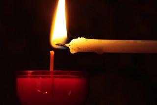 a lit candle is being used to light a tea light candle. most of the image is dark except for the light from the small flame.