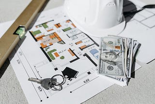 Building plans with some cash, house keys, a level, and a hardhat.