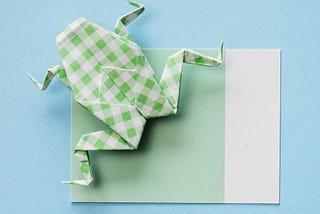 A paper origami frog made of white and green checked back sitting on a piece of green paper with a white strip on one side against a light blue background.