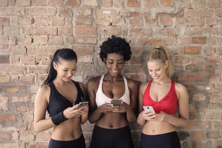 Three women of diverse ethnicity (Asian, African, Caucasian) leaning against an internal brick wall. All three are wearing a sports bra and yoga pants. Each is holding her phone, looking at its screen, and smiling broadly as though amused.