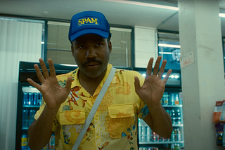 A Black man in a floral shirt and a “Spam” hat has his hands up