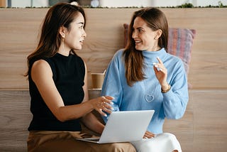 Two women talking to each other about work with a laptop in front of wooden stadium seating