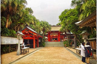 The bright red entrance of a traditional Japanese Shinto Shrine contrasts with the lush greenery surrounding it.