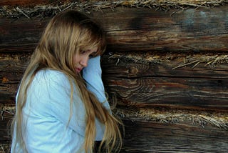 A young woman looks down, leaning against barnwood and hay.