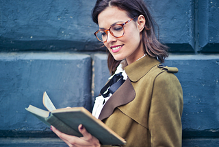 Woman in brown suede peacoat and glasses reading a book