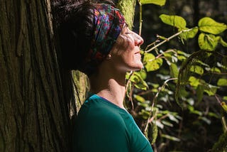 Image of an older woman in a wooded space, leaning against a tree, her eyes closed, with dappled sunlight on her face.