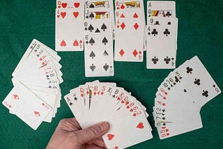 Four hands of cards on a green felt tablecloth — one, at the bottom, is being held by a player; the ones on either side are fanned out, and the one at the top is separated into four piles by suit.