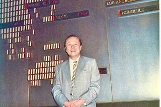 Photo of a smiling middle age man in a suit leaning against a wall chart of global financial reporting.