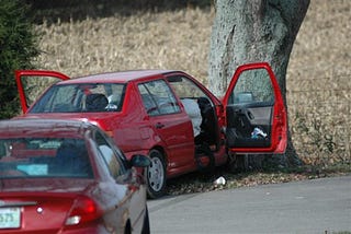 A red car is shown after it has crashed into a treet. Both doors are opened. Another red car is behind it.