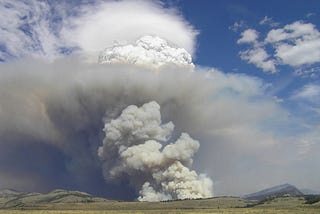 Photograph of a large yet distant cloud of smoke on the horizon. Above and beyond it is a blue cloudy sky. The feeling is of a mind-blowing explosion.