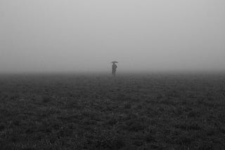 A black & white photo with a person standing on empty land, holding umbrellas, vanishing into the mist.