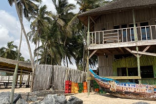 A sraw roofed beach resort with a balcony, surrounded by royal palm trees, blue skies and white clouds. A wooden fence is to the left of the picture and empty soft drink crates. Grey rocks are seen at the foot of the picture, and a canoe boat is in front of the resort with John 3:16 painted onto its side.