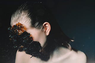 Photograph of a woman. A mass of brown crystals completely obscure her face from the viewer. Her hair is slicked back. The background around her is dark gray.