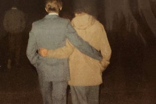 A 16-year-old boy with blonde hair wearing a medium gray suit strolls arm and arm away from the camera with his new lov ein the pitch of night. She has short-cropped black hair and is wearing a khaki jacket.