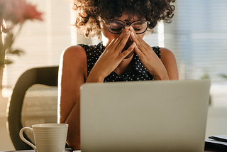 Photo of a woman wearing glasses, looking worried, with their hands on their eyes/nose. A laptop in front of them.