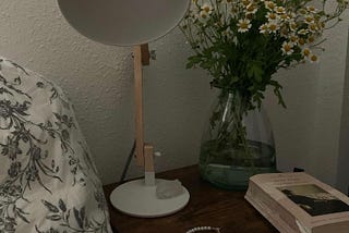 A bedside table, on top of which is a white lamp, chamomile flowers in a vase, a pearl and gold necklace, and a copy of the book “Jane Eyre.”