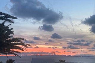 Red sky over Fuerteventura before the big party for Noel’s 80th. It rained all the next day! Dec 2024. Palm trees at one side, the sea in front, and Fuerteventura like a line of bumps, or a low dragon, purple on the horizon, matching the purple cloud in the deepening blue sky.