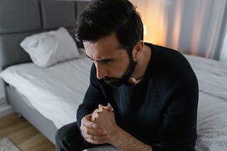 A man in a black shirt sits on an unmade bed.