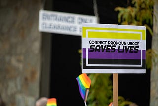A placard is held up with the colours of the non-binary flag and the words, “correct pronoun usage saves lives”.