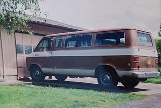 1977 Dodge Van from writer’s childhood, circa 1992, maroon color with white stripes, in front of a tan colored garage door, next to a green lawn, green leaves from tree branches.