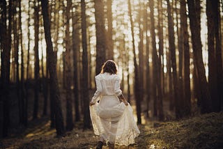 A woman lifts up her white skirt while walking through a forest.