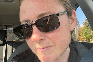 The author has his hair tied back and is sitting in his car, wearing dark sunglasses. He has a serious look on his face and the beginnings of a beard.