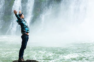 A man stands on a rock surrounded by a river. His arms are out stretched. Behind him, a waterfall rages.