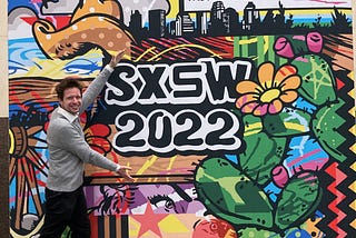 The author poses smiling in front of a colourful sign advertising SXSW 2022, wearing a grey jumper and black trousers.
