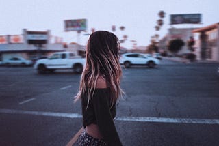 A girl is walking in a city covering her face with her blond hair, few cars are passing on the road