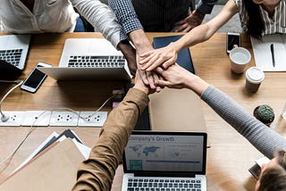 A group puts hands in the middle of the table containing several computers. Crowdsourcing.