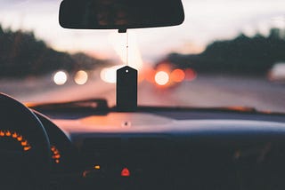 Inside of a car, with a rearview mirror showing. Rt. 66. style. The sun is setting in the background of the highway with trees along both sides.